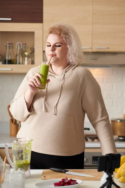 調理ブログを撮影しながら自宅で作ったおいしい緑のスムージーをすすりカービー若い女性 — ストック写真