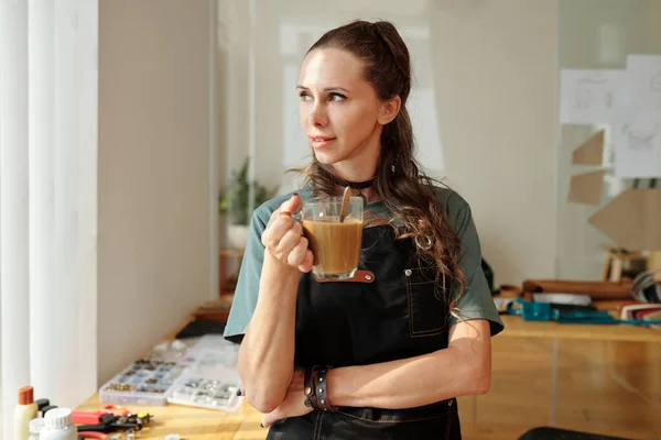 Leather worker drinking coffee in her studio and looking away