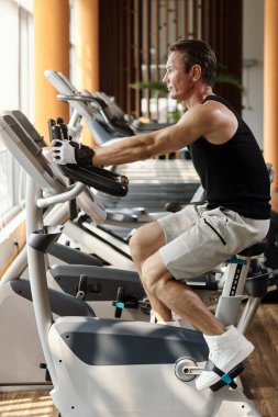 Mature man sitting on exercise bike in gym clipart