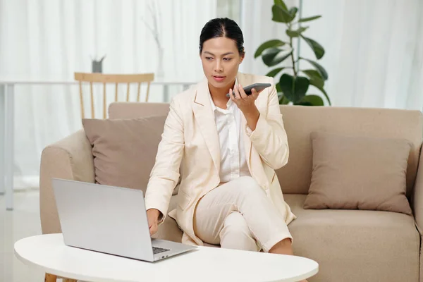 Businesswoman checking e-mails on laptop and recording voice message for colleague