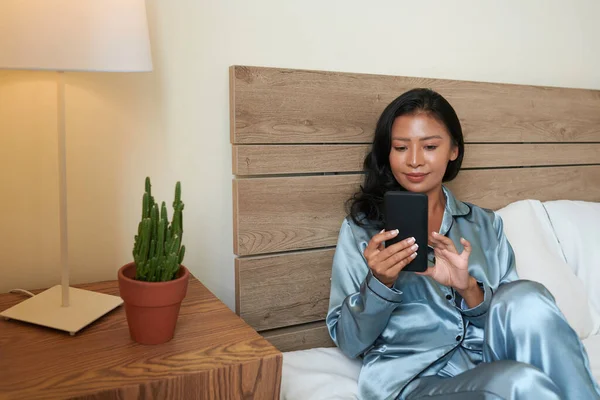 睡衣中的年轻女性坐在床上 查看短信或社交媒体上的帖子 — 图库照片