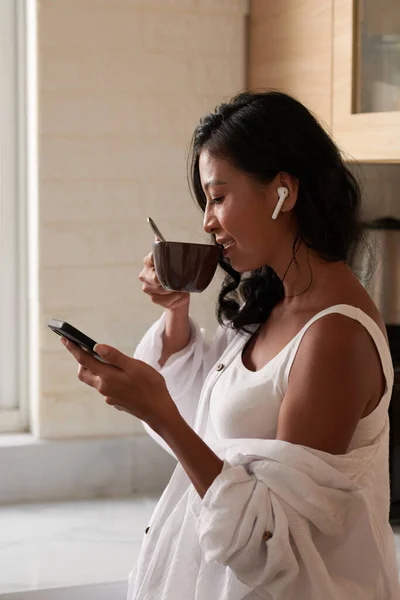年轻女子喝早茶 在智能手机屏幕上阅读文章或文章的侧视图 — 图库照片