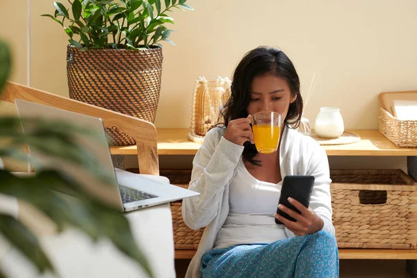 年轻女性在家工作时喝果汁 在智能手机上查看社交媒体 — 图库照片