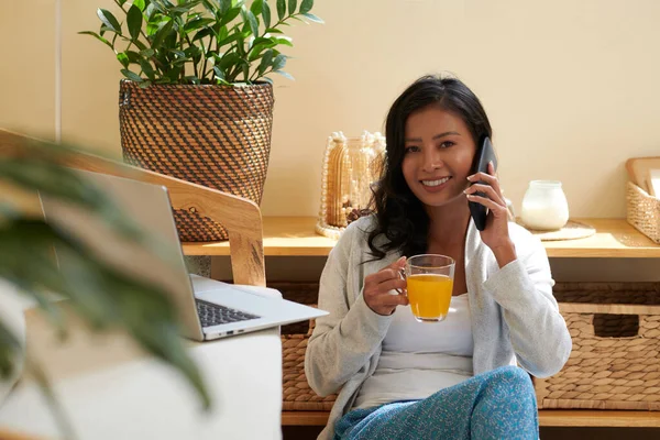 微笑的年轻亚洲女人一边打电话 一边喝着橙汁 — 图库照片