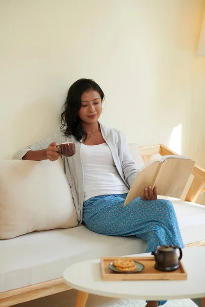 Θετική Νεαρή Γυναίκα Κάθεται Άνετο Καναπέ Πίνοντας Τσάι Και Διαβάζοντας — Φωτογραφία Αρχείου
