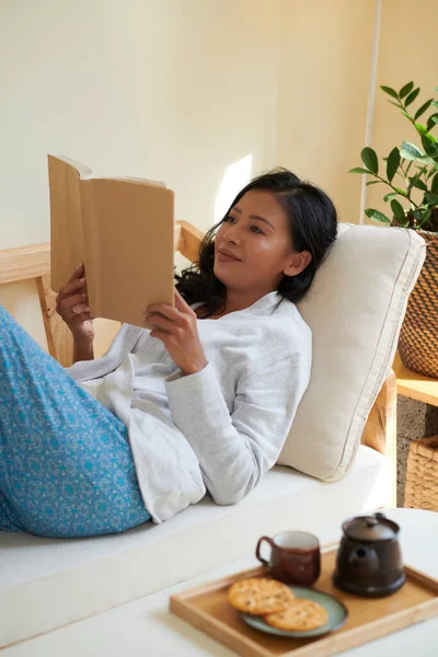 积极的年轻亚洲女人躺在沙发上休息 阅读好书 用饼干喝茶 — 图库照片