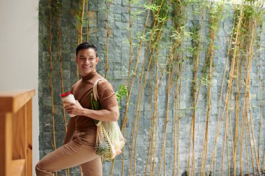 Yeniden kullanılabilir ağ torbası ve açık havada duran tekrar kullanılabilir kahve ile gülümseyen genç adamın portresi.