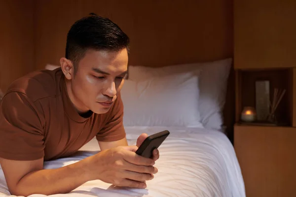 深刻な若い男はベッドの上で休んで 彼の友人からのテキストメッセージに答える — ストック写真