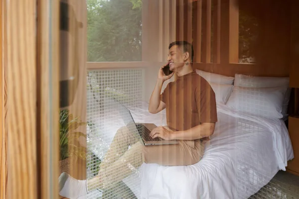 Mutlu Genç Adam Yatakta Oturuyor Arkadaşıyla Telefonda Konuşuyor Dizüstü Bilgisayarında — Stok fotoğraf