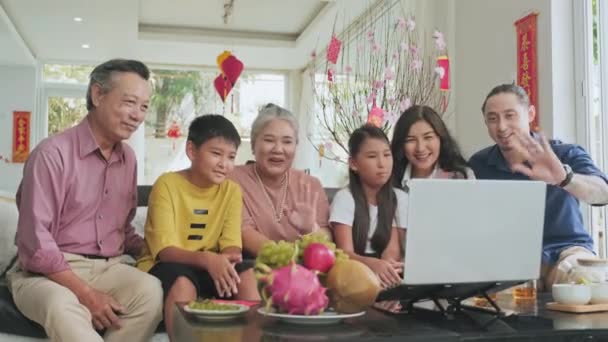 リビングでソファに座っている楽しいアジアの家族は 自宅でTetの休日に集まりながら ラップトップ上のオンラインWebコールを介して親戚と手を振ってチャット — ストック動画