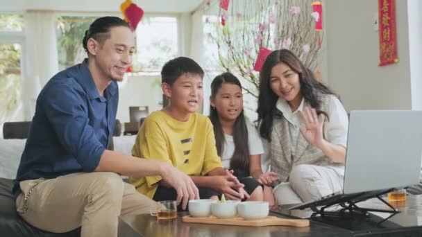 楽しいアジアの家族のカップルと彼らの小さな娘と息子は自宅でソファに座って Tetを祝いながらラップトップでビデオ通話で家族と手を振っておしゃべりします — ストック動画