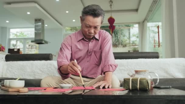 资深亚洲人坐在客厅的沙发上 在准备Tet假日装饰时 用红纸画传统书法 — 图库视频影像