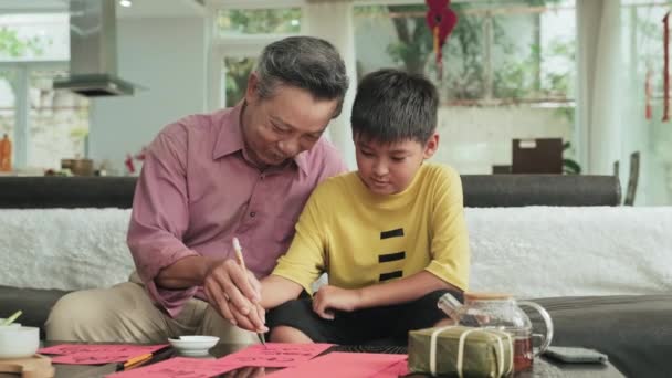 Маленький Азиатский Мальчик Изучает Каллиграфию Веселым Дедушкой Сидя Диване Гостиной — стоковое видео