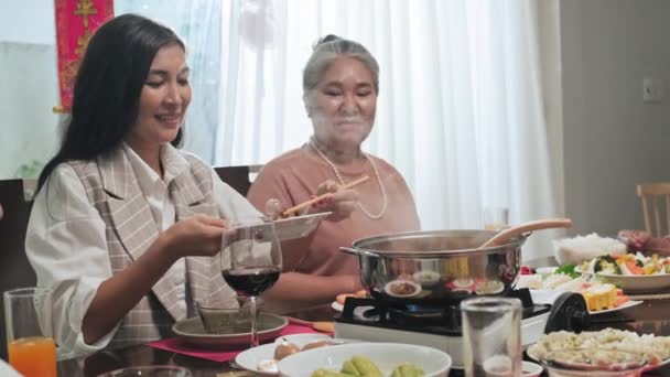 若いアジアの女性はおばあちゃんの隣に座って 家庭でテトの休日の夕食に家族のための伝統的なベトナム料理を作りながら鍋を蒸しに食べ物を入れます — ストック動画