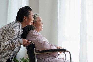 Daughter pushing wheelchair of sick mother to window so she could look outside clipart