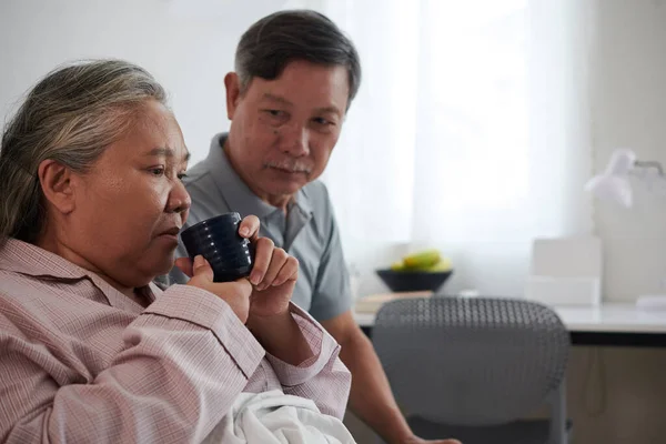 一个严肃的老人看着他生病的妻子在吃药后喝绿茶 — 图库照片