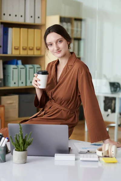 年轻的女商人拿着一杯咖啡坐在写字台上 看着相机 — 图库照片
