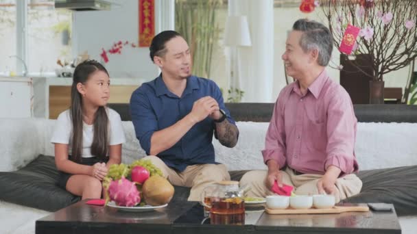 アジア人の家族はソファに座り 老人は小さな孫娘に赤い封筒で幸運を与え 息子は家での旧正月のお祝いのおかげで言う — ストック動画