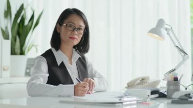 Ofiste evrak işleriyle uğraşırken kameraya gülümseyen genç Asyalı kadın sigortacının orta boy portresi.