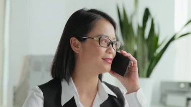 Genç Asyalı kadın yöneticinin göğüs portresi. Telefon görüşmesi yapıyor ve sigorta acentesinde çalışan kameraya bakıyor.
