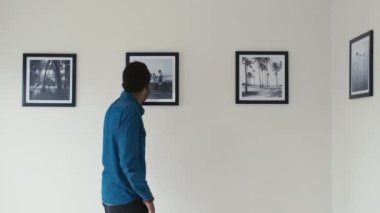 Genç siyahi bir adam sanat galerisinde yürüyor ve sergiyi ziyaret ederken duvarlarda modern fotoğraflar izliyor.