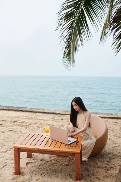 若いです女性でシルクドレスでテーブルに座って熱帯のビーチやラップトップで作業 — ストック写真