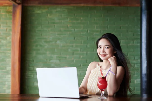 バーカウンターでノートパソコンで働くときにフルーツカクテルを飲む若い女性の笑顔 — ストック写真
