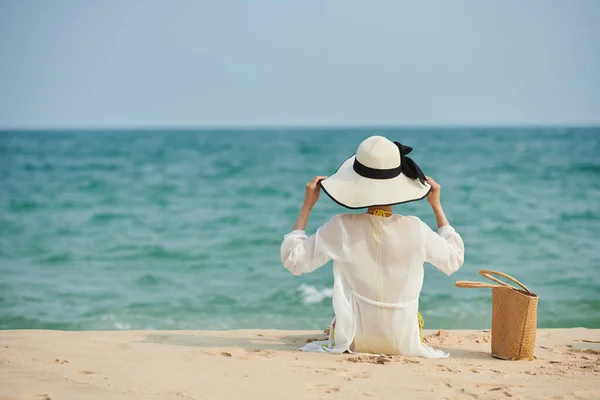 若いです女性に座って砂浜でサテンシャツと帽子と海を見て — ストック写真
