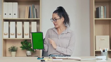 Kadının kameraya bakıp dijital tablette uygulamayı yeşil renkli ekranla sunarken orta boy görüntüsü.