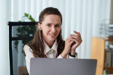 İş yerinde bilgisayarla otururken kameraya gülümseyen kulaklıklı genç bir iş kadınının portresi