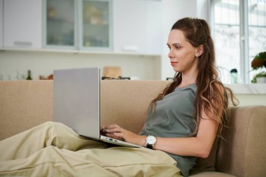 Kanepede oturan ve boş zamanlarında dizüstü bilgisayar kullanan genç bir kadın.