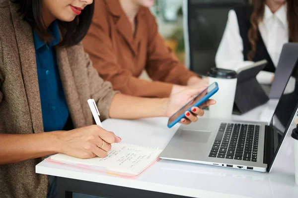 Zbliżenie Businesswoman Pomocą Smartfona Notatek Notatniku Podczas Pracy Laptopem — Zdjęcie stockowe
