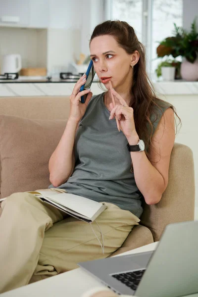 ソファにメモ帳を置きながら携帯電話で話している若い女性の垂直イメージ 彼女は自宅で働いています — ストック写真