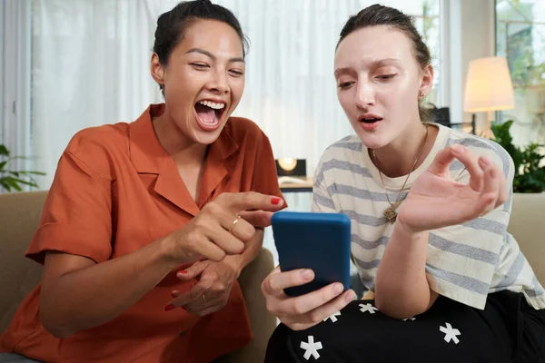 两个朋友坐在客厅的沙发上 一边用智能手机看滑稽的照片 — 图库照片