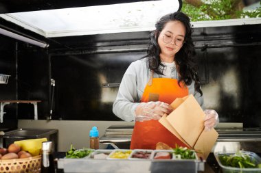 Üniformalı ve eldivenli genç bayan tezgahtar fast food yemek için kese kağıdı tutarken sokak arabasının müşterileri için sosisli sandviç paketliyor.