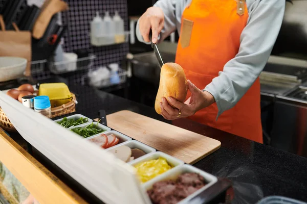 お客様のためにホットドッグを準備しながら 木製のまな板上に新鮮なパンを切断オレンジ色のエプロンの若い食品業者のクローズアップ — ストック写真