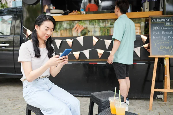 年轻的亚洲女孩坐在街上的快餐车旁 一边用智能手机打滚 一边等男朋友买热狗或汉堡包 — 图库照片