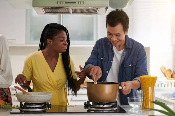 年轻的跨文化夫妇穿着便装 在煤气炉上准备意大利面 而黑人妇女则拿着煎锅帮助她的亚洲男友 — 图库照片