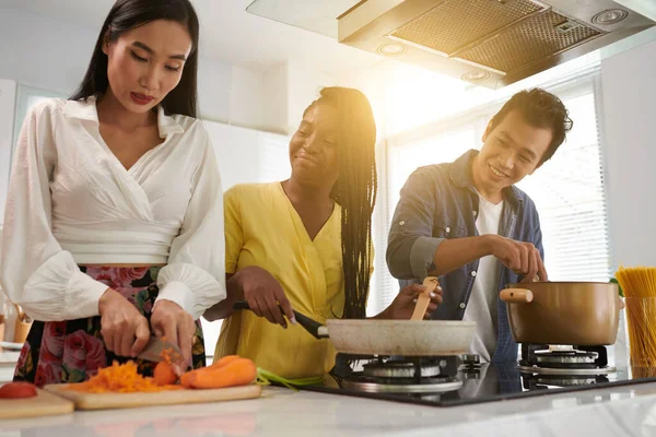 台所のガスストーブのそばに立っている若い黒人女性 フライパンで食材を混合し 彼女の友人を見て新鮮なニンジンをみじん切り — ストック写真