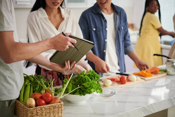 若い男は台所のテーブルのそばに立って彼の妻にタブレット画面上でオンラインレシピを示し 友人と野菜サラダや他の料理を準備 — ストック写真