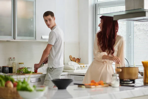 身穿T恤衫的年轻人站在厨房柜台边 一边看他的妻子 一边讨论晚餐的菜单 一边切蔬菜做沙拉 — 图库照片