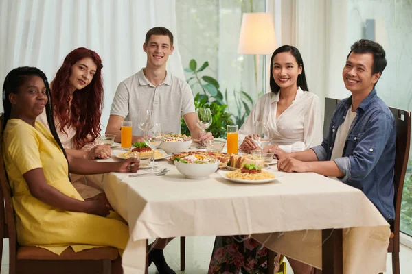 五位年轻的多元文化朋友坐在餐桌旁看着相机 端着自制的开胃菜 开胃菜和橙汁杯 — 图库照片
