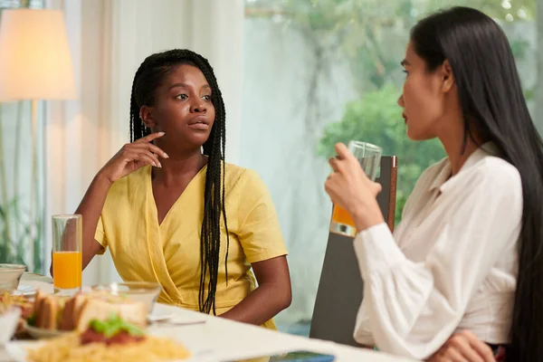 身穿黄色衣服的年轻的非洲裔美国女人一边坐在餐桌旁聊天 一边看着那位喝着果汁的亚洲朋友 — 图库照片