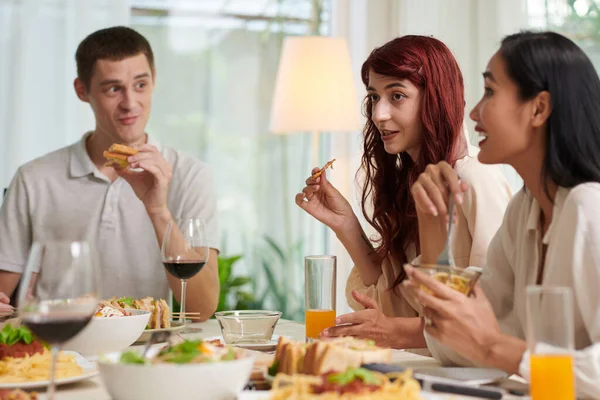夕食中にテーブルに座り おいしい自家製の食べ物を食べ 何かを議論することで彼女の友人の間に座って軽食を持つ若い女性 — ストック写真