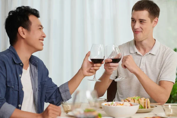 テーブルに座りながら赤ワインを飲みながら二人の若い異文化間の仲間が手作りの食べ物を提供し 夕食を楽しむ — ストック写真