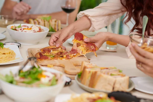 若い空腹の女性の手料理と夕食のテーブルの上に立って木製の板から食欲をそそる自家製ピザのスライスを取ります — ストック写真
