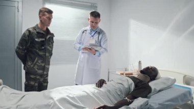 Beyaz üniformalı profesyonel bir doktor hastanede askerle birlikte yaralı bir hastanın teşhisini tartışıyor.