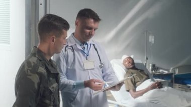 Beyaz önlüklü doktor ve üniformalı asker askeri hastanenin koğuşunda yatan yaralı bir hastanın tedavisinden bahsediyorlar.