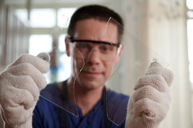 Koruyucu eldivenler giymiş bir zanaatkar elinde iki cam parçasıyla camdan şekil kesiyor.