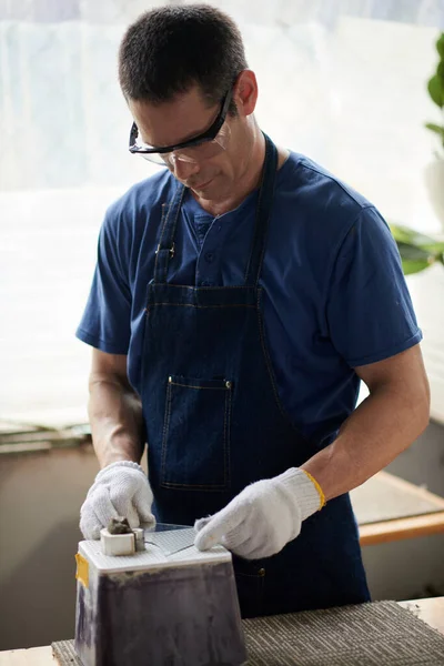 Vertical image of worker in protective gloves and glasses making artwork from glass in workshop